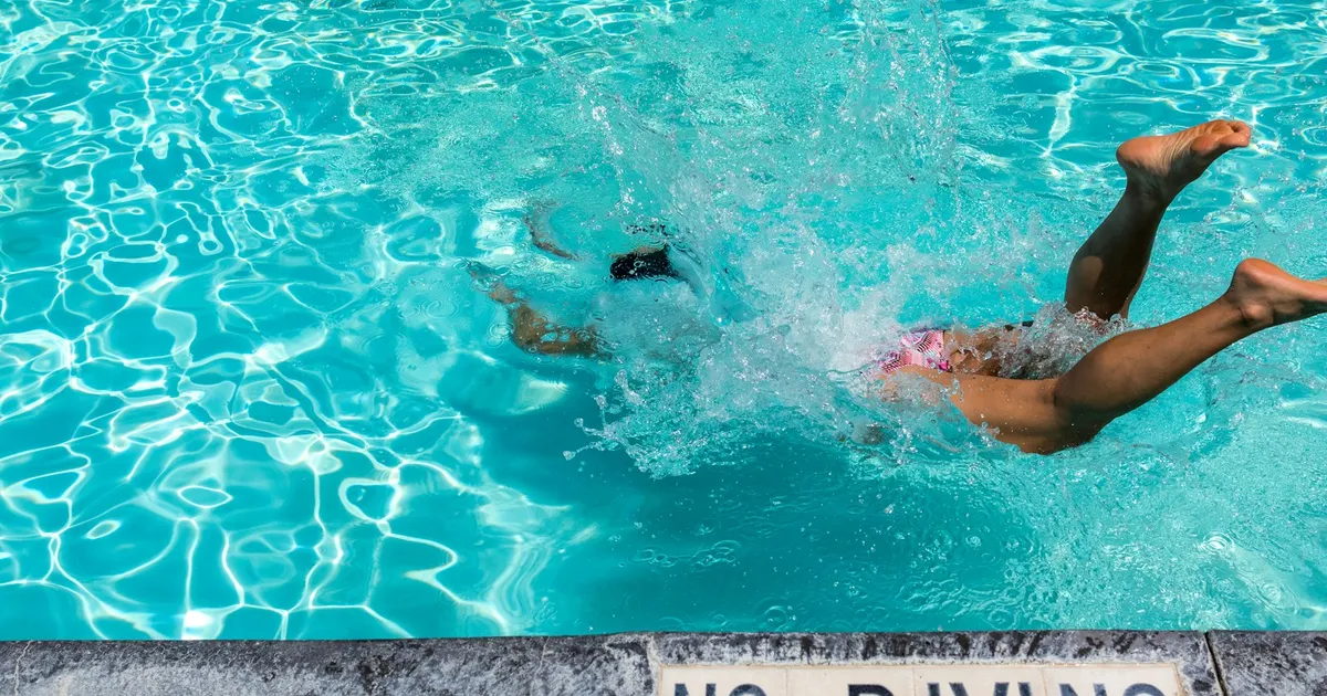 Iemand duikt met een plons in een helderblauw zwembad naast een No Diving-tegel
